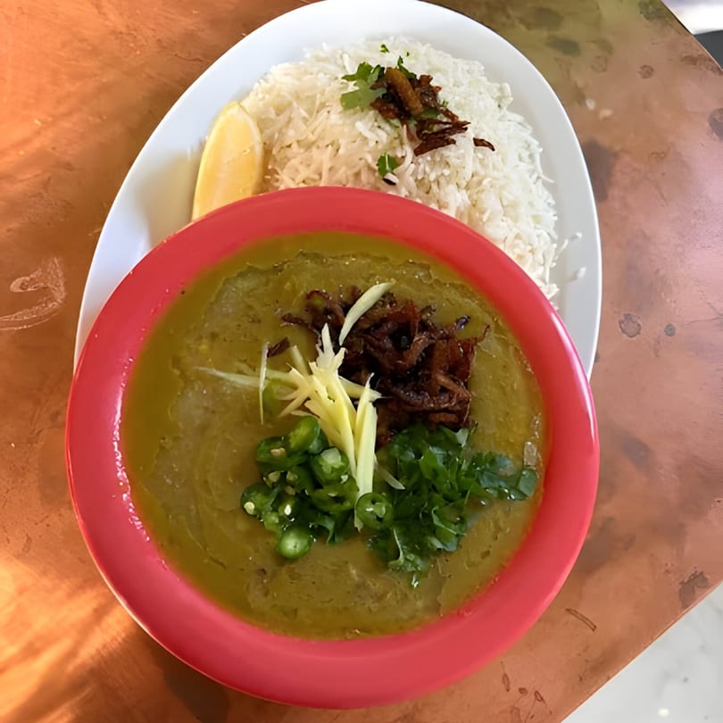 Pakistani haleem in a red bowl with rice, lemon, and fried onions on a white plate.